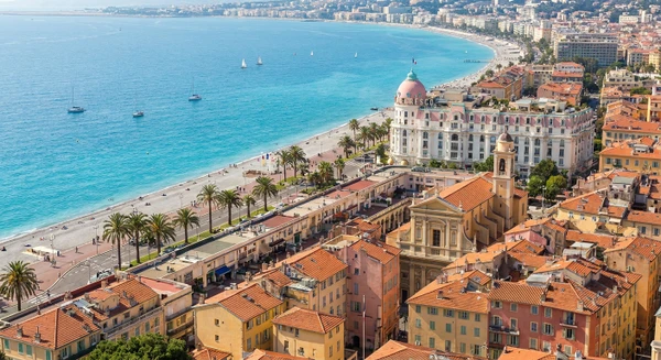 Photographie réaliste de la ville de Nice, France, montrant des éléments architecturaux et urbains caractéristiques : vue sur la Promenade des Anglais avec ses bâtiments emblématiques, ou une perspective des quartiers reconnaissables de Nice (vieille ville avec ses façades colorées, front de mer, ou environnement urbain côtier typique). La photo doit être prise de jour, bien éclairée, avec une qualité professionnelle, sans texte ni logo visible. Elle doit représenter distinctement Nice (non simplement une ville côtière générique) pour se démarquer visuellement des images existantes pour Cannes et Antibes.